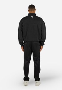 Sort sweatshirt med høj krave og elastiske manchetter, parret med sorte sweats, der har et hvidt logo på ryggen og hvide sko.
