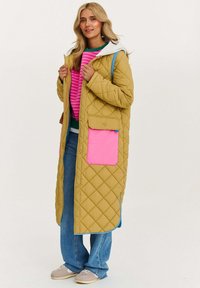 Prešívaný dlhý kabát v horčicovej žltej farbe s pink vreckom, šedou kapucňou a modrými akcentmi. Nosený cez sveter s ružovými pruhmi a modré džínsy.