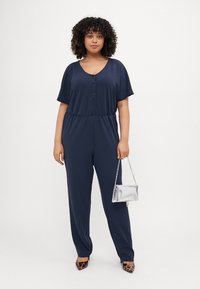 Navy blue jumpsuit with short sleeves and button details, elastic waistband, paired with a silver clutch and leopard print heels.