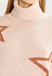 Ljusrosa stickad tröja med hög polokrage och en framträdande rustfärgad stjärnmotiv. Mjuk textur med ribbdetaljer.