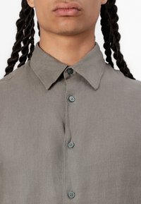 Camisa de lino gris con cuello puntiagudo, que presenta botones azul claro y una textura visible, mostrada desde el pecho hacia arriba.