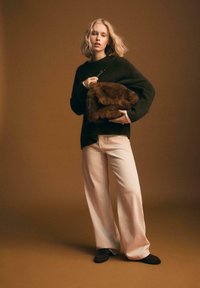 Donna che indossa un maglione nero e pantaloni chiari, mentre tiene una grande borsa di pelliccia marrone su uno sfondo marrone uniforme.
