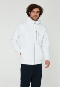 Bílá mikina na zip s kapucí se stahovací šnůrkou, obsahující náprsní kapsu na zip a žebrované manžety, nošená přes tmavé kalhoty.