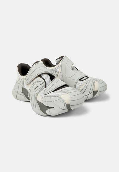 Paire de baskets futuristes blanches et grises avec des surpiqûres en caoutchouc texturé et des brides en Velcro, posées sur un fond blanc uni.