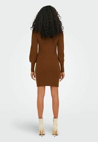 Robe en tricot côtelé marron à manches longues et silhouette ajustée, se terminant à mi-cuisse. Associée à des bottines crème à talons hauts.