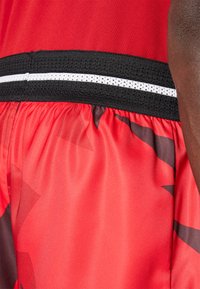 Rote sportliche Shorts mit glatter Textur, ausgestattet mit einem schwarzen elastischen Bund mit einem weißen Streifen und subtilen dunklen geometrischen Mustern.