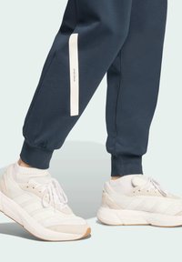 Námornícke tepláky s bielym bočným pruhom a textom "SPORTSWEAR", spárované s bielymi teniskami s béžovými detailmi a gumenou podrážkou.