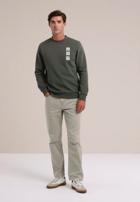 Hombre de pie con una mano en el bolsillo, vistiendo un sudadera verde oscuro con diseños abstractos en blanco, jeans gris claro y zapatillas blancas.