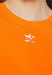 Oranžové bavlněné tričko s kulatým výstřihem a žebrovaným lemem, na kterém je malé bílé logo "adidas" s třemi listovými detaily.