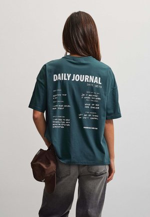 Persona vista desde atrás que lleva una camisa verde oscuro con texto impreso de diario y sostiene una gorra marrón en una mano.