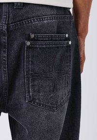 Vaqueros de mezclilla negra con un bolsillo trasero que presenta un detalle de costura en forma de estrella y remaches de metal, mostrando una tela texturizada.