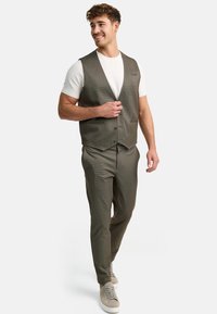 Olivenkariertes Weste über einem weißen T-Shirt, kombiniert mit passenden karierten Hosen und hellgrauen Sneakers. Lässiger, taillierter Schnitt.