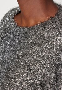 Gros plan du cou et des épaules portant un pull texturé argenté brillant avec des fibres métalliques et un col arrondi.