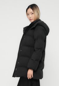Frau mit hellbraunem Haar, die eine schwarze gepolsterte Kapuzenjacke trägt, seitlich vor einem schlichten hellen Hintergrund steht.
