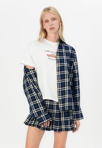 White t-shirt with printed text, layered under a blue and black plaid shirt. Matching plaid skirt with pleats. Smooth texture, casual design.