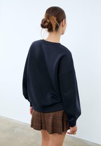Felpa blu navy oversize con scollo a giro e spalle cadenti, abbinata a una mini gonna plissettata a quadri marrone, vista da dietro.