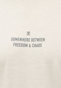 Κείμενο «Κάπου ανάμεσα στην ελευθερία και το χάος» με γκρι γράμματα σε ανοιχτό μπεζ υφασμάτινο φόντο, με μικρό αφηρημένο σύμβολο πάνω από το κείμενο.