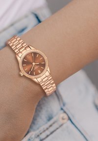 Roosi kuldne kell ümmarguse näo, pruuni gradueeritud tausta ja kuldsete tunni markeritega. Omab lingitud metallist rihma ja tekstureeritud serva.