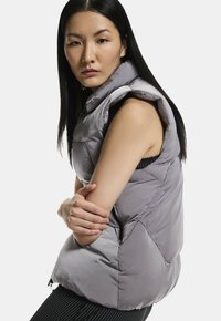 Grå puffervest med høj krave, ærmeløs design og en tekstureret overflade. Har lynlås lukning og geometriske syninger.