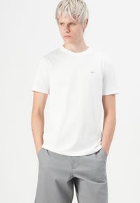 Junger Mann mit kurzen blonden Haaren, der ein schlichtes weißes T-Shirt und graue Hosen trägt, mit den Händen in den Taschen steht und nach links schaut.