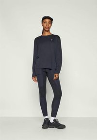Top nero a maniche lunghe con scollatura tonda e logo, abbinato a leggings neri con pannelli testurizzati. Il modello indossa sneakers nere e un berretto.