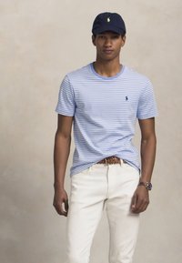 Jaunas vyras su tamsiai mėlyna kepure, šviesiai mėlynu raištiniu marškinėliais, baltais kelnais, rudais pintais diržais ir rankiniu laikrodžiu, stovintis prieš neutralų foną.