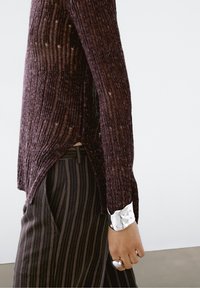 Pull en maille bordeaux avec texture côtelée, fentes latérales et poignets contrastants blancs. Associé à un pantalon marine à fines rayures et des accessoires en argent.