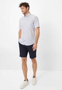 Camicia a maniche corte con bottoni di colore azzurro chiaro con un motivo geometrico, abbinata a pantaloni corti blu scuro e sneaker bianche.