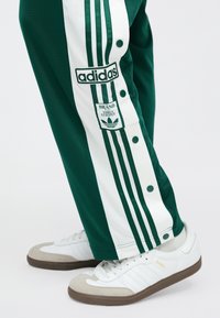 Πράσινο παντελόνι φόρμας με λευκές ρίγες στο πλάι, με κουμπιά τύπου σναπ και λογότυπο Adidas. Κλασικά λευκά αθλητικά παπούτσια με γκρι και χρυσές λεπτομέρειες.