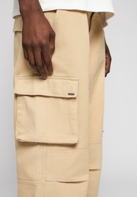 Pantalon cargo beige en tissu texturé, avec une poche à rabat sur le devant et des détails de couture apparents. Une main repose près de la poche.