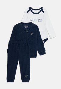 Marineblaues, strukturiertes Jacken- und Hosen-Set mit einem passenden weißen Langarm-Bodysuit, der mit marineblauen Akzenten und dem „GUESS“-Logo in mehrfarbigen Buchstaben versehen ist.