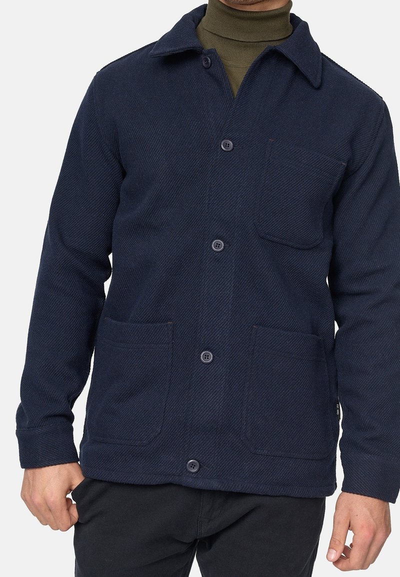 Homme portant une veste bleu marine boutonnée avec trois poches avant sur un pull à col roulé vert olive et un pantalon noir.