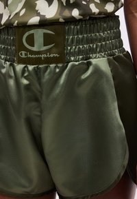Σκούρο πράσινο σατέν αθλητικό σορτς με φαρδιά ελαστική μέση που φέρει λογότυπο Champion, φορεμένο με τοπ καμουφλάζ.