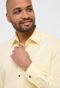 Camicia leggera gialla con un sottile motivo a righe, con polsini abbottonati e taschino sul petto. Bottoni a contrasto ben visibili sul colletto e sui polsini.