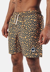 Navy blue swim shorts with a repeating orange elephant pattern, elastic waistband, and drawstring. Features a circular logo patch on the side.