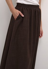 Jupe maxi brune, texturée, avec taille élastique et poches latérales ; son design fluide améliore le tombé et le mouvement.