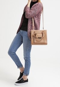 Béžová ramenní taška s přední kapsou, hladká kožená textura. Modelka má na sobě růžový vzorovaný cardigan, černý top a vyhrnuté modré džíny s černými slip-on botami.