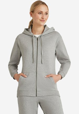 Vrouw draagt een lichtgrijze hoodie met ritssluiting en trekkoorden, samen met bijpassende trainingsbroek, handen in de zakken, kijkt lichtjes opzij met een glimlach.