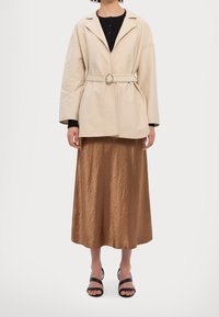 Beige Jacke mit Gürtel und weiten Ärmeln, getragen über einem schwarzen gerippten Top und einem glänzenden braunen Midirock. Schwarze Riemchensandalen vervollständigen das Outfit.