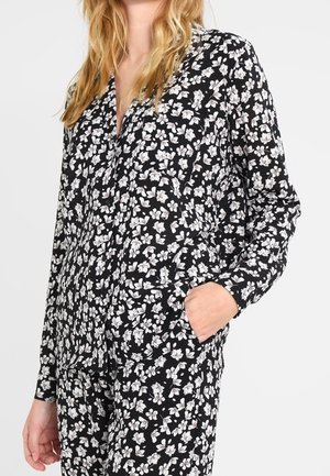 Kvinde iført sort skjorte med knapper og lange ærmer samt bukser med lille hvidt blomsterprint, med den ene hånd i lommen, stående foran en ensfarvet baggrund.