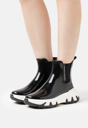 Bottes de pluie noires brillantes à la cheville avec de lourdes semelles blanches et noires, portées sur des jambes nues devant un fond uni.