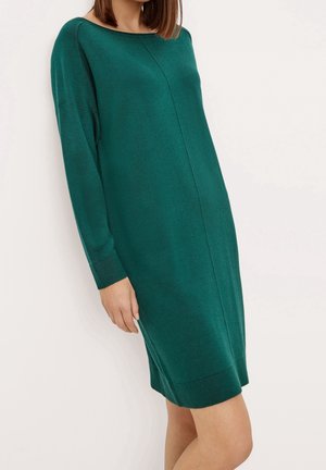 Femme portant une robe en maille vert foncé, longue jusqu'aux genoux, manches longues, avec un décolleté bateau, debout devant un fond clair uni.