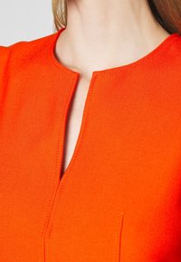 Vêtement orange avec un col montant doté d'une ouverture en forme de goutte, texture lisse et couture précise le long des bords.