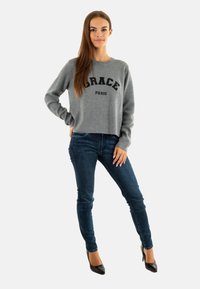 Maglione grigio corto con la scritta "GRACE PARIS" in bold nero, abbinato a jeans skinny blu scuro e tacchi a punta neri. Texture liscia.