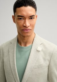 Hellgrauer, strukturierten Blazer mit Reverskragen, versehen mit einer weißen Blumenbrosche, über einem mintgrünen Rundhalsausschnitt-T-Shirt. Glatter Stoff und moderner Schnitt.