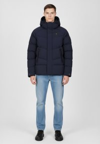 Granatowa kurtka puchowa z kapturem, wyposażona w zapinane na zamek boczne kieszenie, pikowany wzór i wysoki kołnierz, zestawiona z jasnoniebieskimi dżinsami i czarnymi butami.