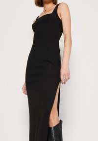 Robe noire sans manches avec une silhouette ajustée, dotée d'une fente sur le côté, réalisée en tissu lisse, mettant en avant un design minimaliste.