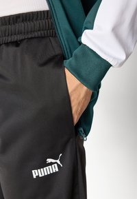 Svart trening bukser med en glidelåslomme, elastisk midjebånd og hvit Puma-logo. Den tilhørende jakken er petrolfarget med hvite ermer.