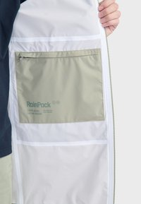 Ľahká pršiplášť s bielym exteriérom, so zipsom upevnenou olivovo zelenou kapsou s nápisom "RainPack" vo vnútri. Obsahuje podšívku a akcenty.