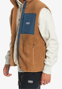 Brun fleeceväst med blå nylonbröstficka, hel dragkedja, sidofickor och en mjuk textur. Lägger sig över en grå sweatshirt.